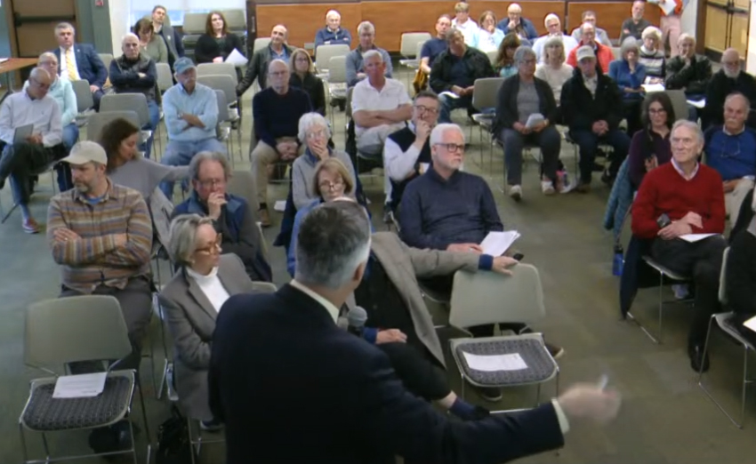Several dozen seated attendees listening to the presenter in the foreground.